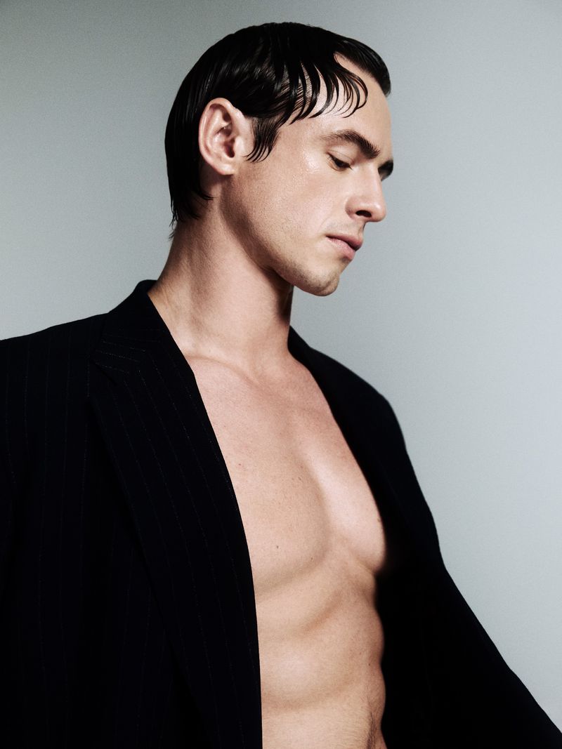 Headshot of a man with slicked-back dark hair wearing an open black blazer, gazing down in a photo studio with soft, neutral lighting.