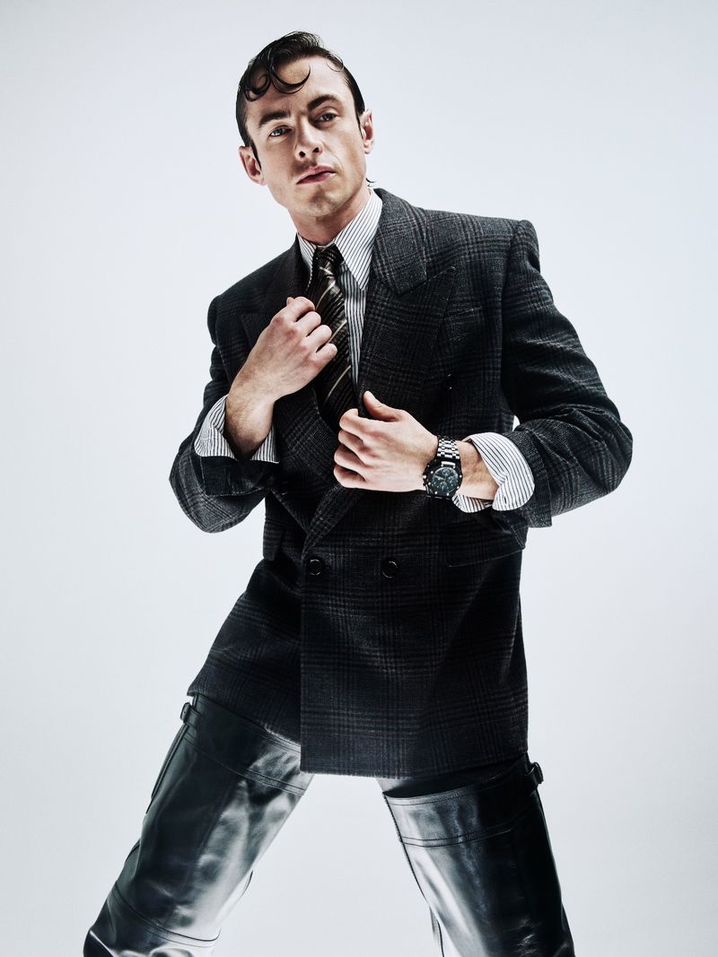Studio portrait of a man in a suit jacket and riding boots striking a pose against a white background, with dark hair slicked back.