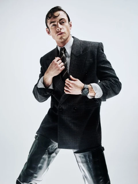 Studio portrait of a man in a suit jacket and riding boots striking a pose against a white background, with dark hair slicked back.