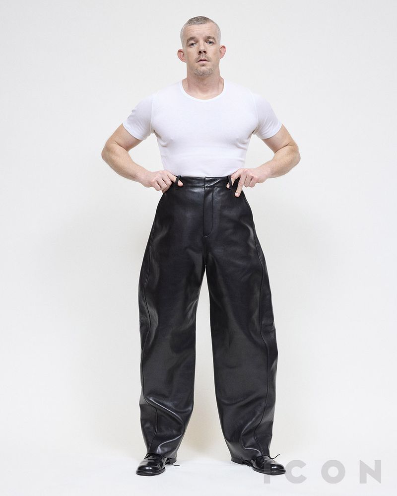 Man in white t-shirt and oversized black leather pants standing in a light studio. Full shot of adult male fashion.