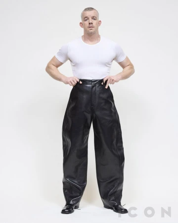 Man in white t-shirt and oversized black leather pants standing in a light studio. Full shot of adult male fashion.