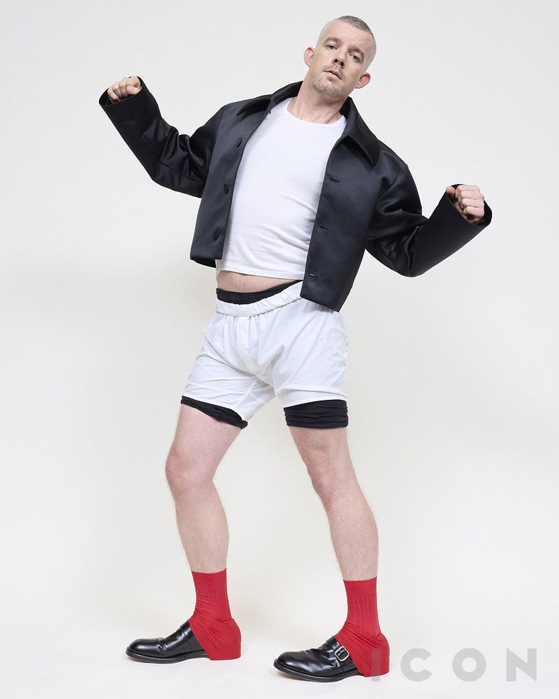 Full shot of adult male in black jacket, white t-shirt, shorts, red socks, and dress shoes posing in a photo studio against white backdrop.
