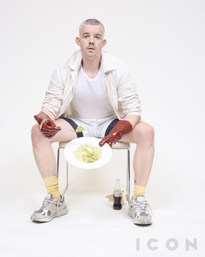 Man in open jacket and shorts sits holding plate of greens with large gloves on in a studio with a spilled soda.