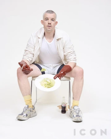 Man in open jacket and shorts sits holding plate of greens with large gloves on in a studio with a spilled soda.