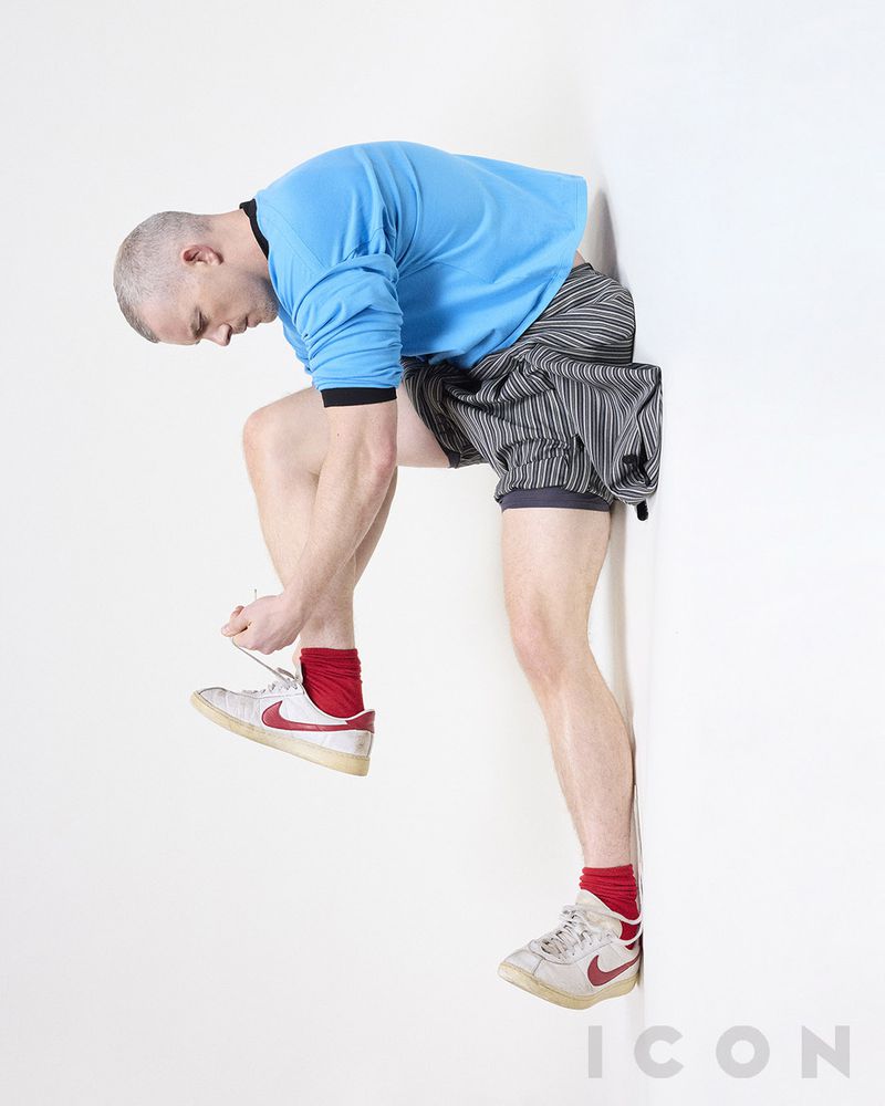 Athletic bald man in blue shirt ties shoelaces on vintage sneakers against a white wall, creating a studio portrait.