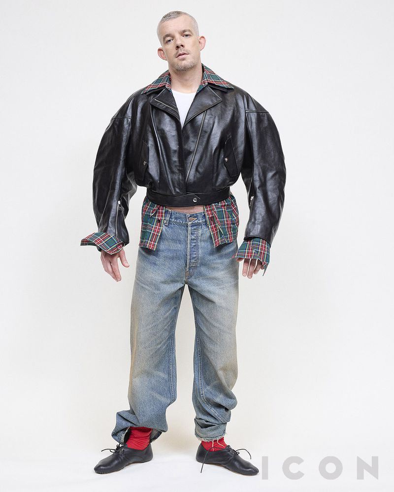 Adult male poses indoors wearing oversized leather jacket, t-shirt, baggy jeans, socks, and dress shoes against a white background.