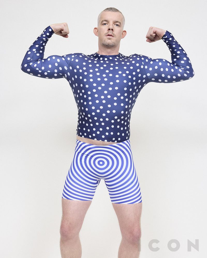 Athletic build man flexing in a photo studio. He is wearing a blue polka dot top and blue swirl patterned shorts against a white backdrop.