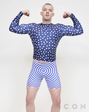 Athletic build man flexing in a photo studio. He is wearing a blue polka dot top and blue swirl patterned shorts against a white backdrop.