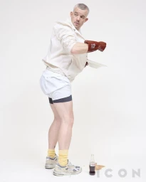 Actor Russell Tovey wearing an off-white jacket, athletic shorts, rubber gloves, and yellow socks against a white studio backdrop.