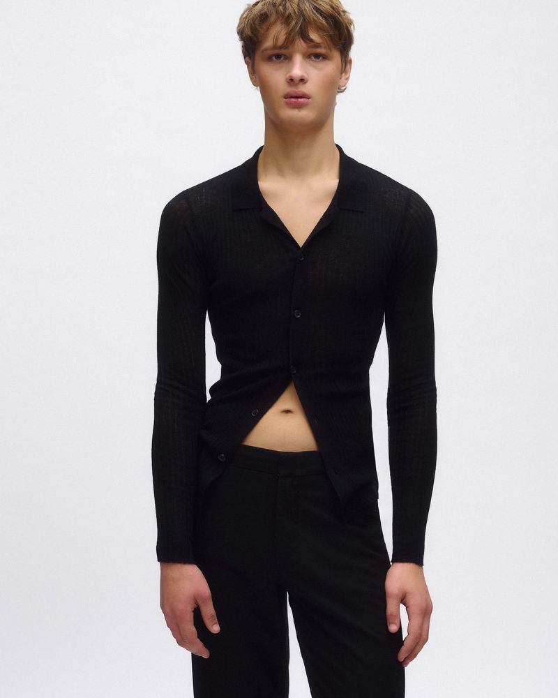 Fashion portrait of a young adult male wearing a black, buttoned long-sleeve shirt with black pants posing against a white backdrop.