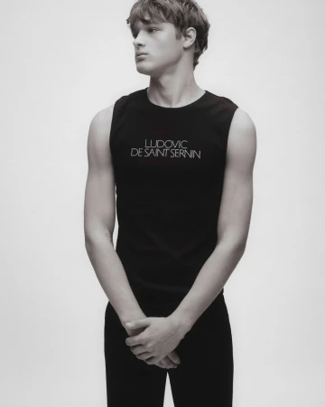 Studio shot of a young adult male in black tank top and black pants, arms folded, looking left in a high-key setting with soft lighting.