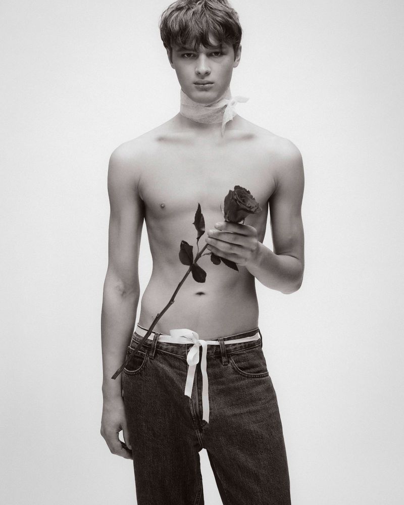 Portrait of a young shirtless man holding a single rose, wearing jeans and a white sash around his neck, set against a clean, white background.