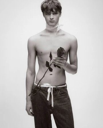 Portrait of a young shirtless man holding a single rose, wearing jeans and a white sash around his neck, set against a clean, white background.