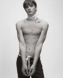 Monochrome portrait of a shirtless young man wearing a white gauze neck wrap and dark jeans against a studio backdrop.