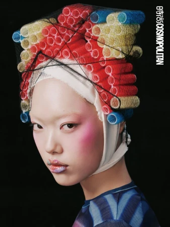 Asian female fashion model poses in photo studio with curlers on her head, pink blush and artistic lipstick against black background.