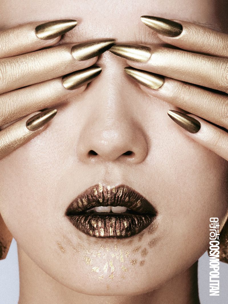 Close-up of a woman with golden skin, nails, and lips, covering her eyes with her hands, creating a striking and artistic beauty shot.