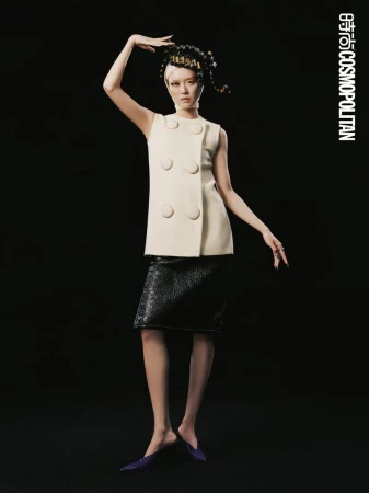A single woman poses in a sleeveless white dress and black skirt against a black background in a studio with the Cosmopolitan logo.