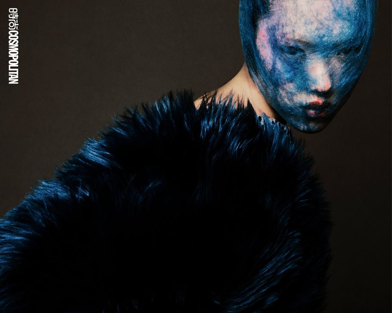 Close-up studio shot of a model wearing a textured full-face mask, and a large dark faux fur coat, against a dark background.