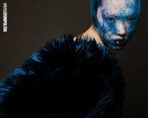 Close-up studio shot of a model wearing a textured full-face mask, and a large dark faux fur coat, against a dark background.