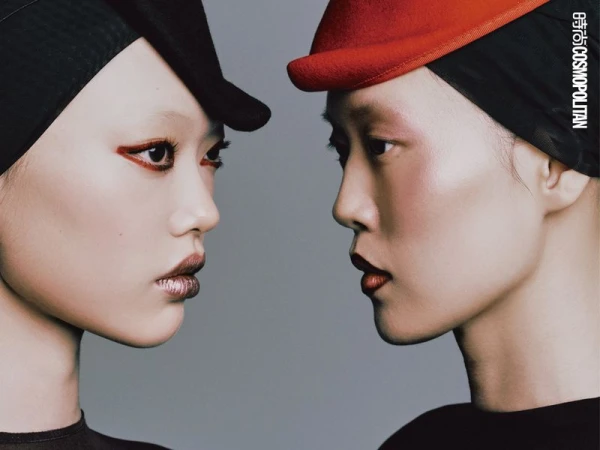 Close-up profile portrait of two women, one wearing a black cap, the other a red cap, both with stylized makeup on a gray background.