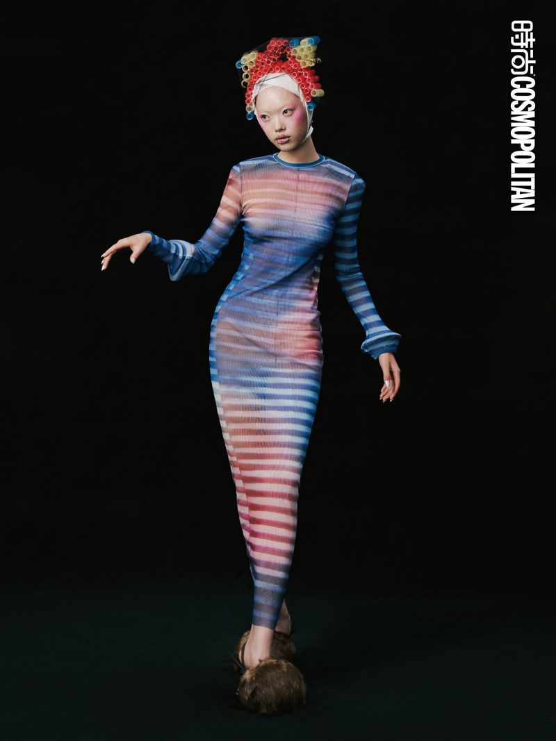 Fashion editorial shot: a slender woman in a striped dress poses against a dark backdrop, showcasing avant-garde hair and statement shoes.