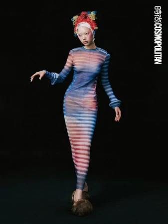 Fashion editorial shot: a slender woman in a striped dress poses against a dark backdrop, showcasing avant-garde hair and statement shoes.