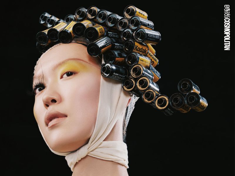 Asian woman with film roll hair and yellow eyeshadow against black background. Cosmopolitan logo visible, fashion beauty shoot.