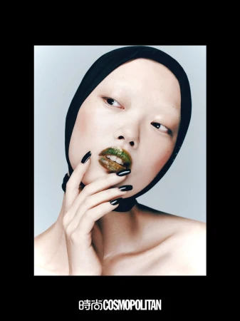 Close-up beauty shot of an Asian woman wearing a black head covering and green and gold lip gloss, posing in a photo studio.