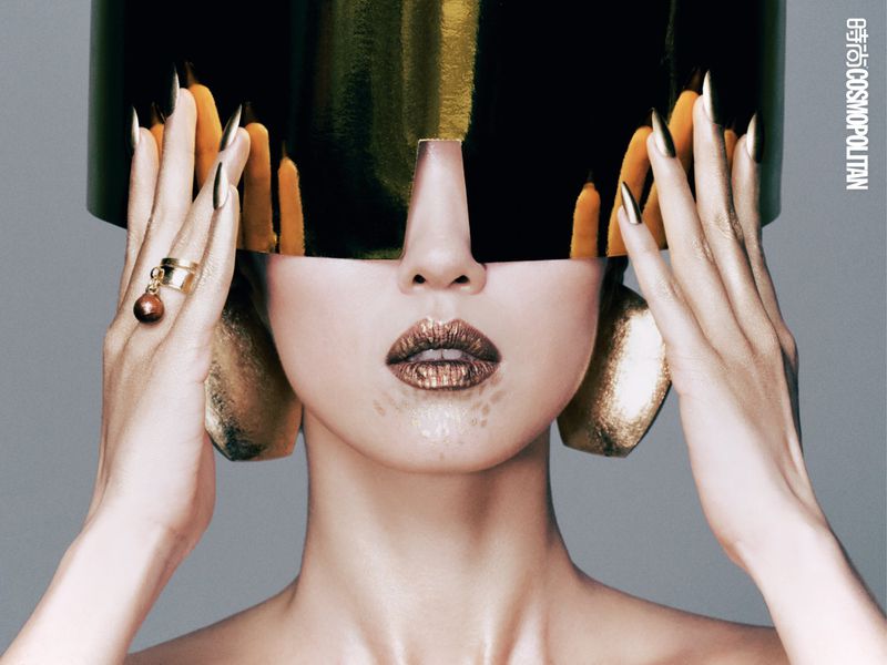 Close-up of a woman with artistic makeup, wearing a golden helmet-like headpiece, gold jewelry, and gold fingernails against a gray background.