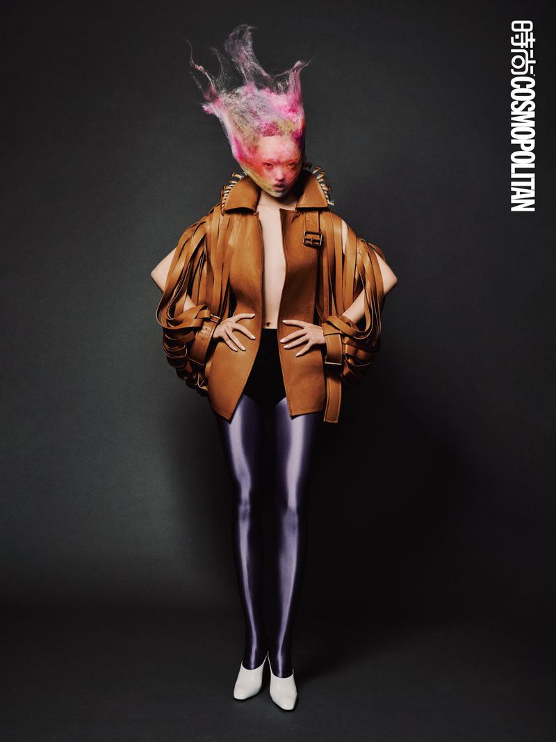 Fashion shot of an adult female wearing a tan jacket with shoulder pads, black leggings, and white heels against a gray backdrop.
