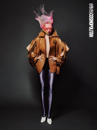 Fashion shot of an adult female wearing a tan jacket with shoulder pads, black leggings, and white heels against a gray backdrop.