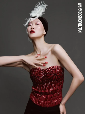 Elegant woman in strapless top, black head wrap with white feather, and bright red lipstick and nail polish poses in studio.