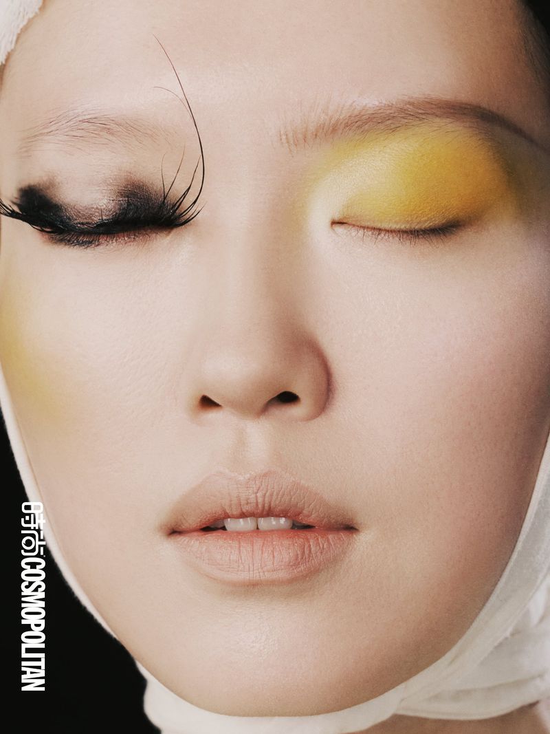 Close-up of an Asian woman's face with dramatic eye makeup featuring oversized eyelashes on one eye and yellow eyeshadow on the other.