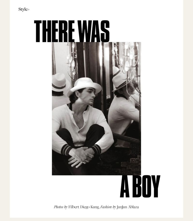 Black and white portrait of a fair-skinned adult male in a white hat and sweater, seated with knees up, in a fashion editorial.