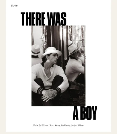 Black and white portrait of a fair-skinned adult male in a white hat and sweater, seated with knees up, in a fashion editorial.