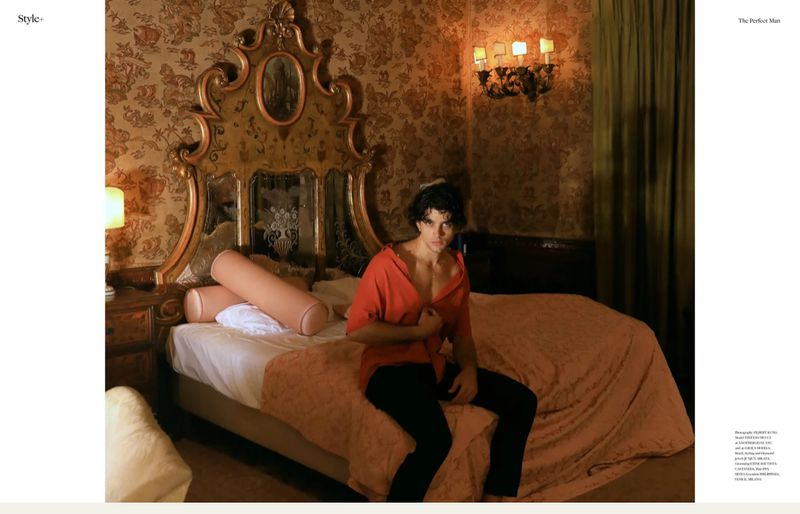 Fashion shot of a young adult male with dark hair sitting on the edge of a bed in a room with floral wallpaper and ornate headboard.