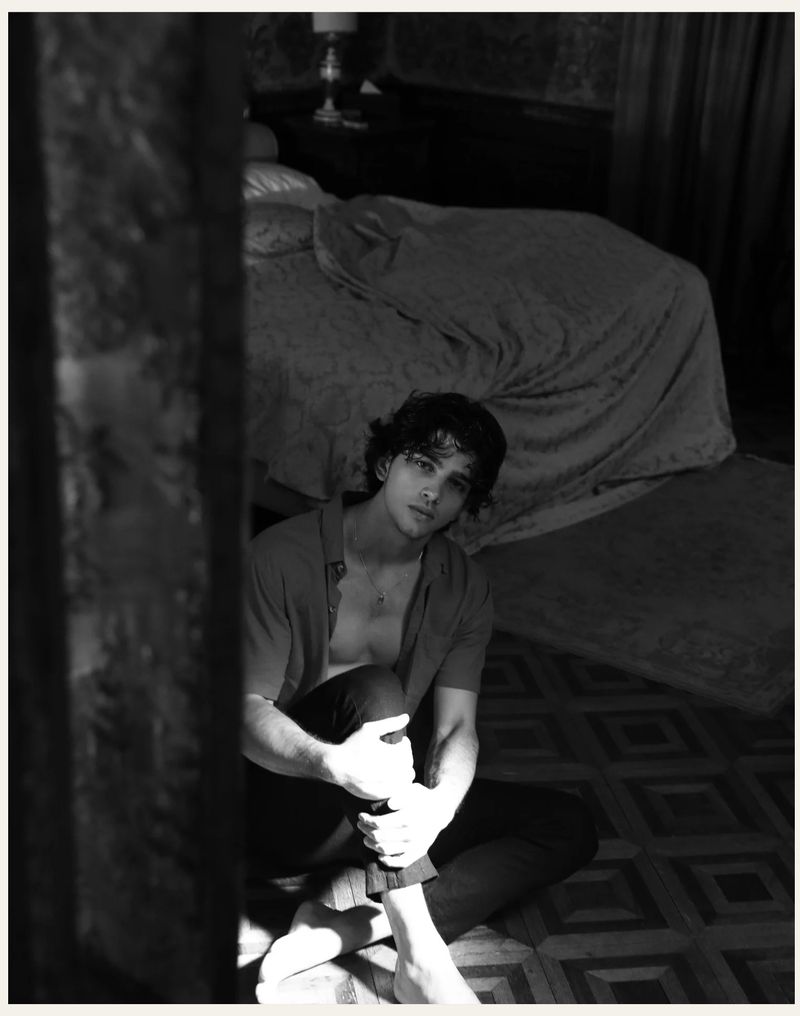 Black and white portrait of a young adult man with curly hair, sitting barefoot on a patterned floor in a bedroom, looking at the camera.