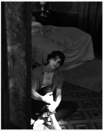 Black and white portrait of a young adult man with curly hair, sitting barefoot on a patterned floor in a bedroom, looking at the camera.