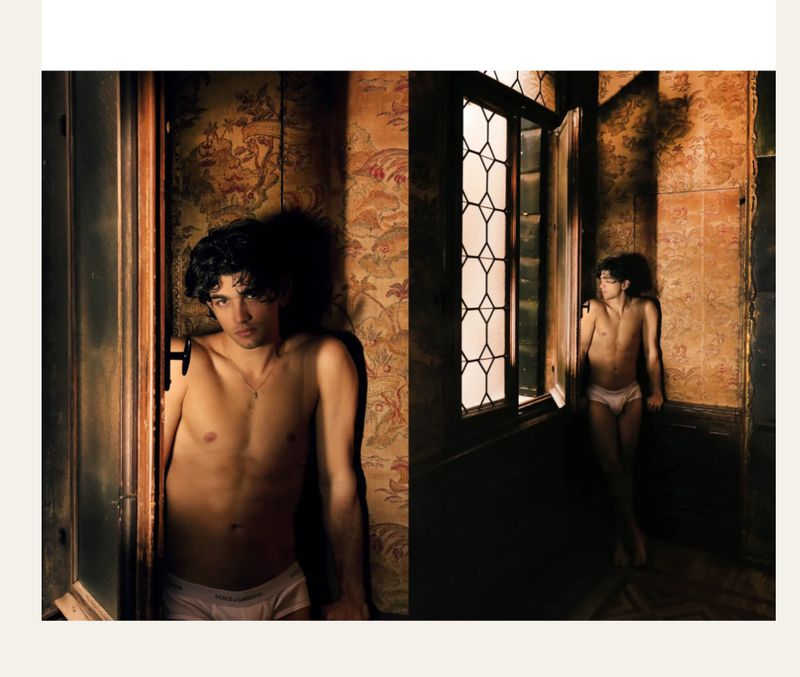 Striking indoor fashion shot features a shirtless, lean young man in briefs looking toward a window with patterned wallpaper.