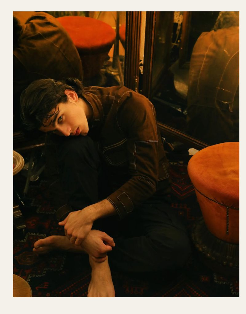 Young man in dark shirt sitting cross-legged on a patterned rug near orange stools reflected in mirrors