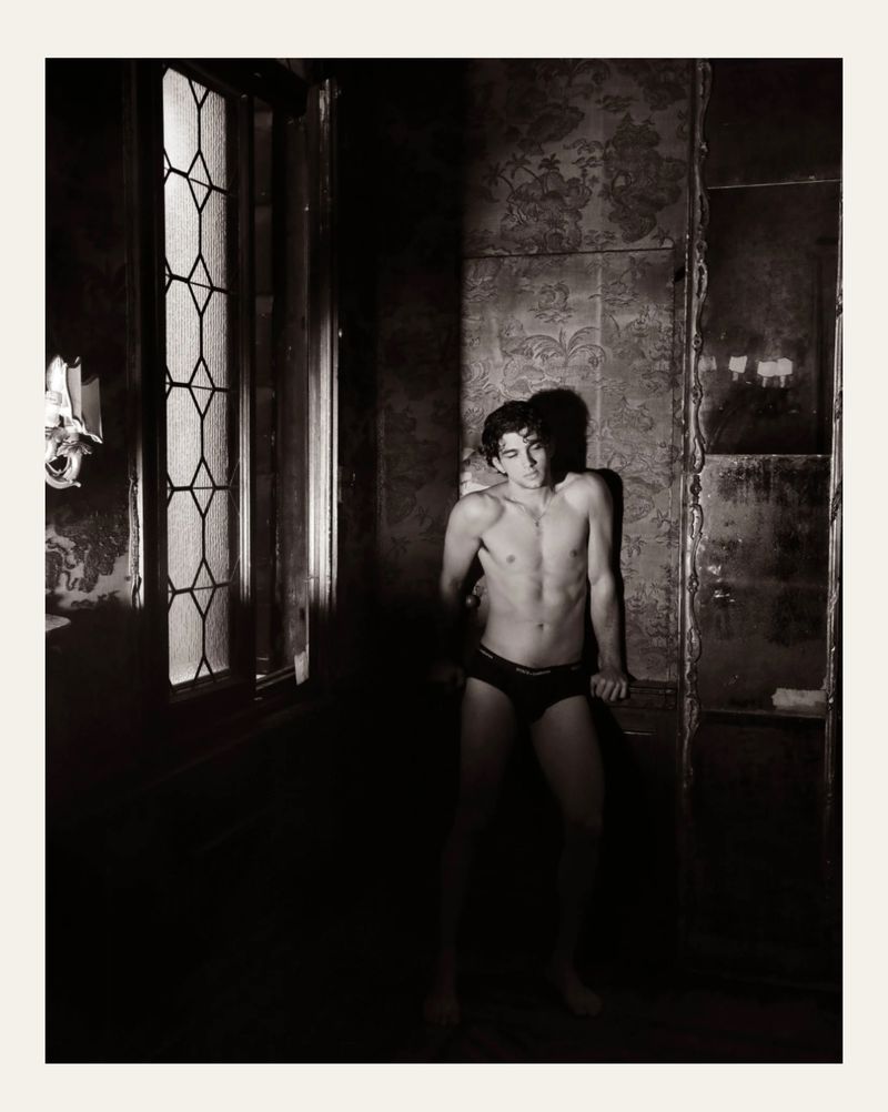 Monochrome portrait of a lean adult male in underwear posing in dimly lit, vintage-style room with ornate wallpaper and a window.