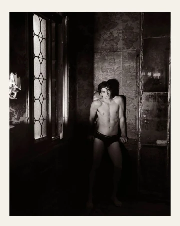 Monochrome portrait of a lean adult male in underwear posing in dimly lit, vintage-style room with ornate wallpaper and a window.