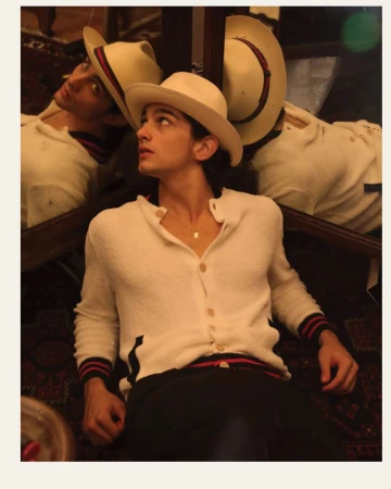 Portrait of a young man wearing a cowboy hat and cardigan, looking up. Reflected in a mirror with a patterned background.