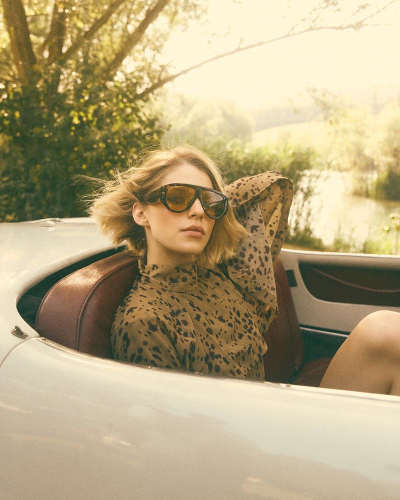 Model Amaëlle Gery wearing oversized tortoiseshell sunglasses and a patterned blouse in a vintage white convertible car.