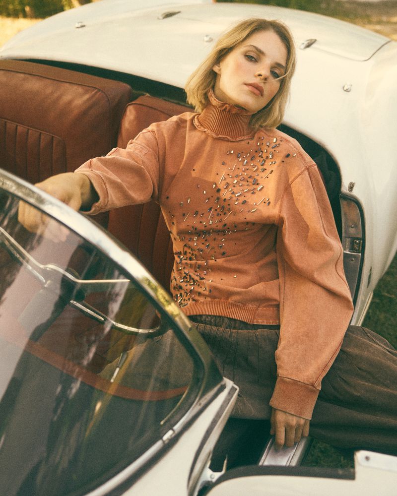 Fashion shot of a fair-skinned young adult woman with short blonde hair sitting in a vintage convertible car wearing a rust-colored long-sleeve sweater.