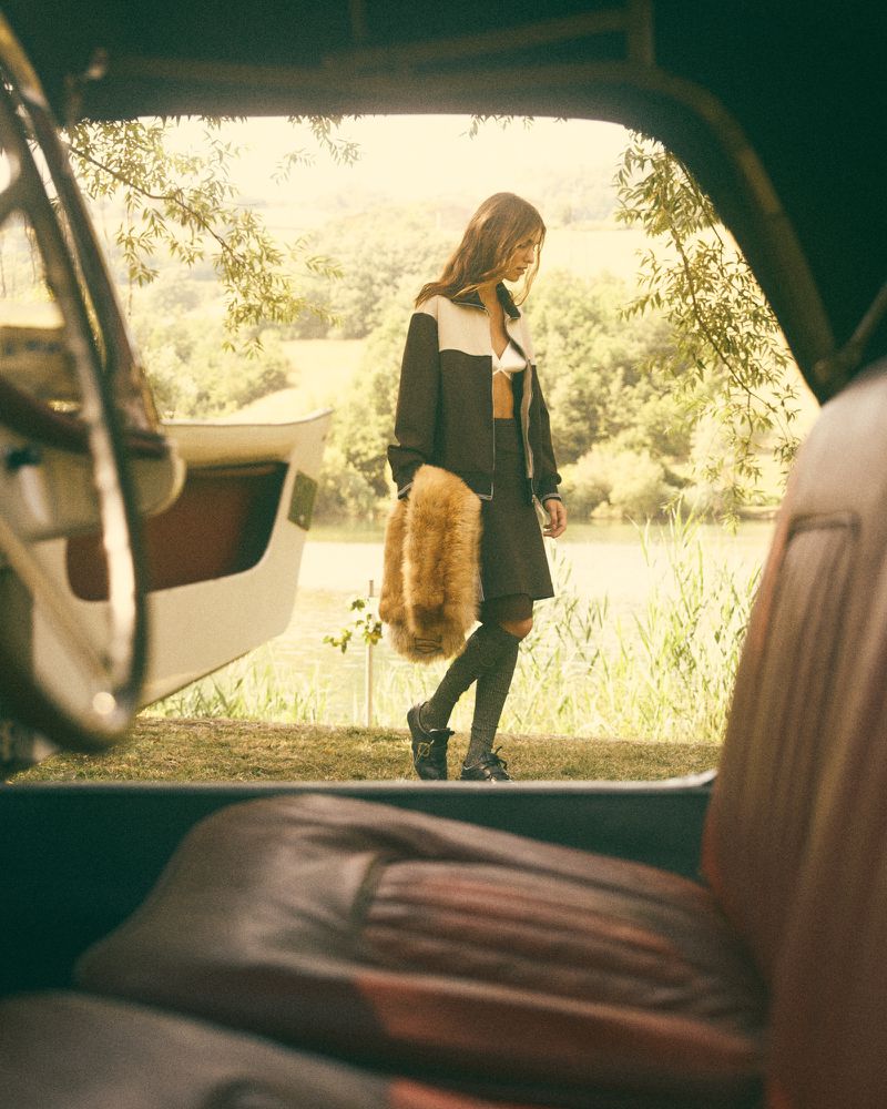 A woman in a jacket and skirt walks near a classic car with a river and trees in the background on a sunny day.
