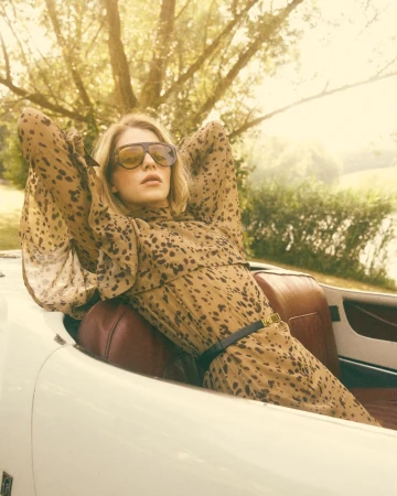 A blonde woman in a patterned dress and sunglasses reclines in a vintage white convertible car with a soft-focus outdoor backdrop.