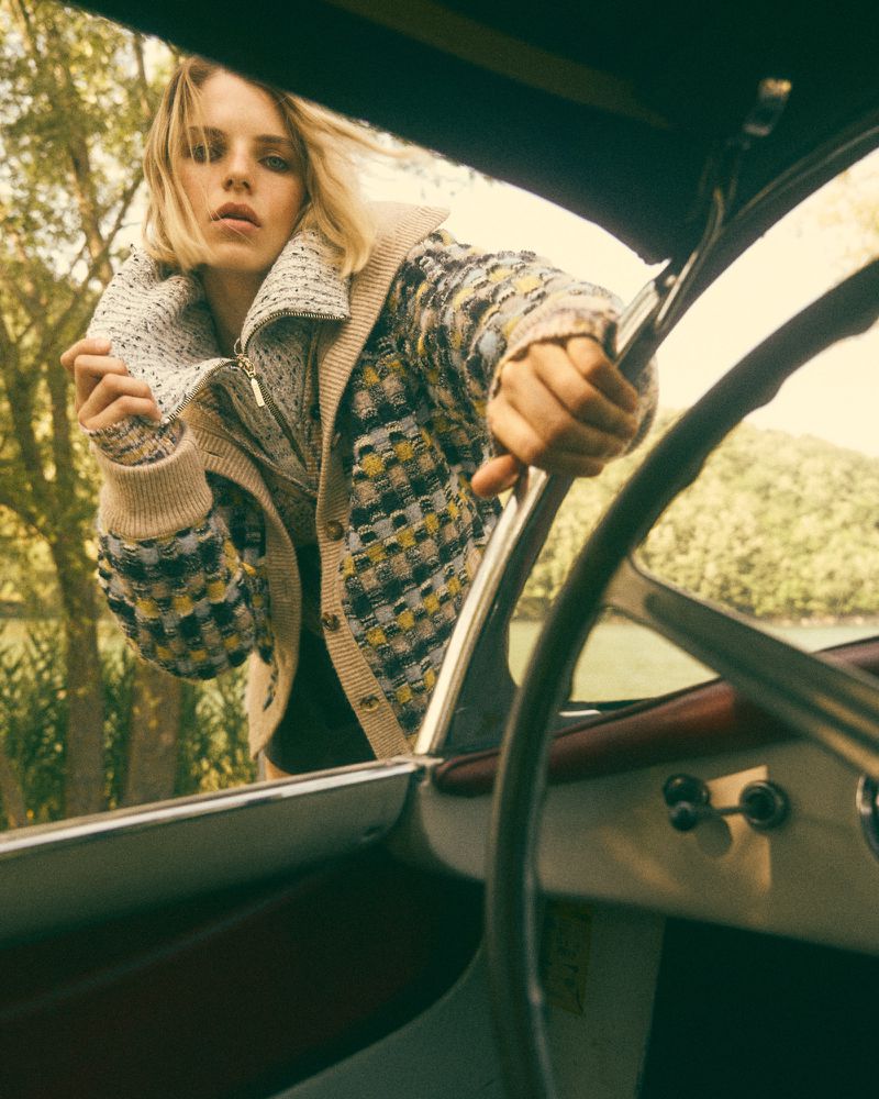 A blonde woman in a patterned cardigan sits in a boat, holding the steering wheel, with trees and water visible in the background.