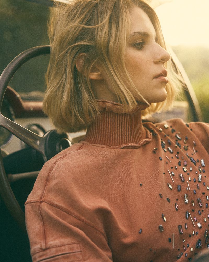 Woman in rusty-orange embellished sweatshirt sits in a vintage car with a steering wheel, side-profile view, soft sunlight, outdoor.