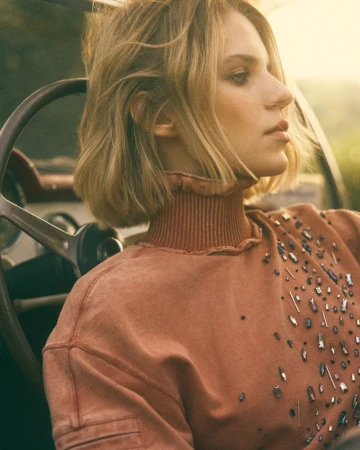 Woman in rusty-orange embellished sweatshirt sits in a vintage car with a steering wheel, side-profile view, soft sunlight, outdoor.